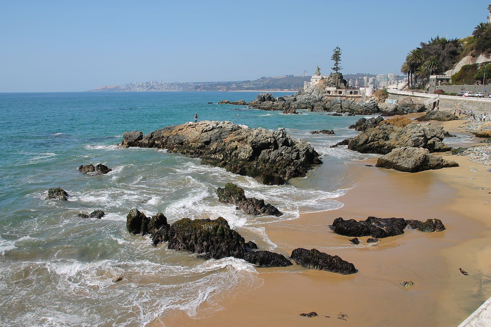 Felsen und goldener Sand an der Uferpromenade von Viña del Mar, mit dem Castillo Wulff auf der Landspitze und der Bucht dahinter